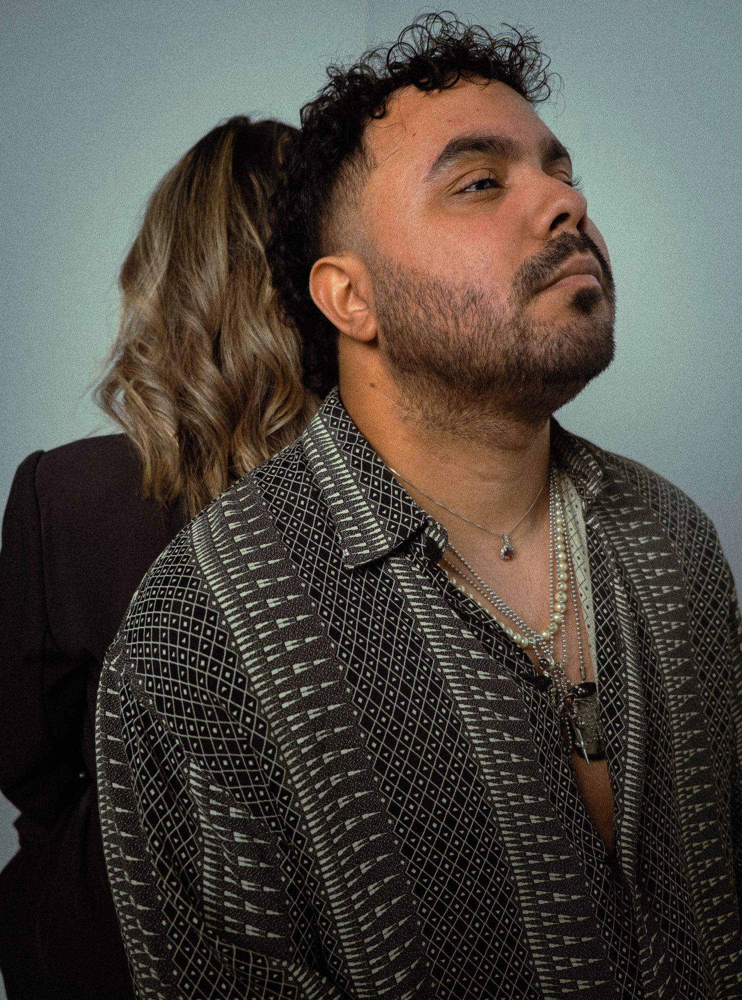 Groom-to-be in a patterned shirt, his fiancée behind him on a sage studio backdrop